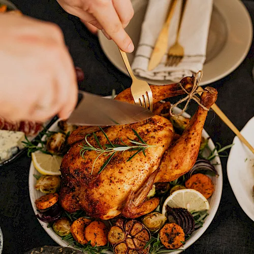 A cook is carving a roasted chicken with a fork and knife, serving it on a platter with vegetables and lemon.