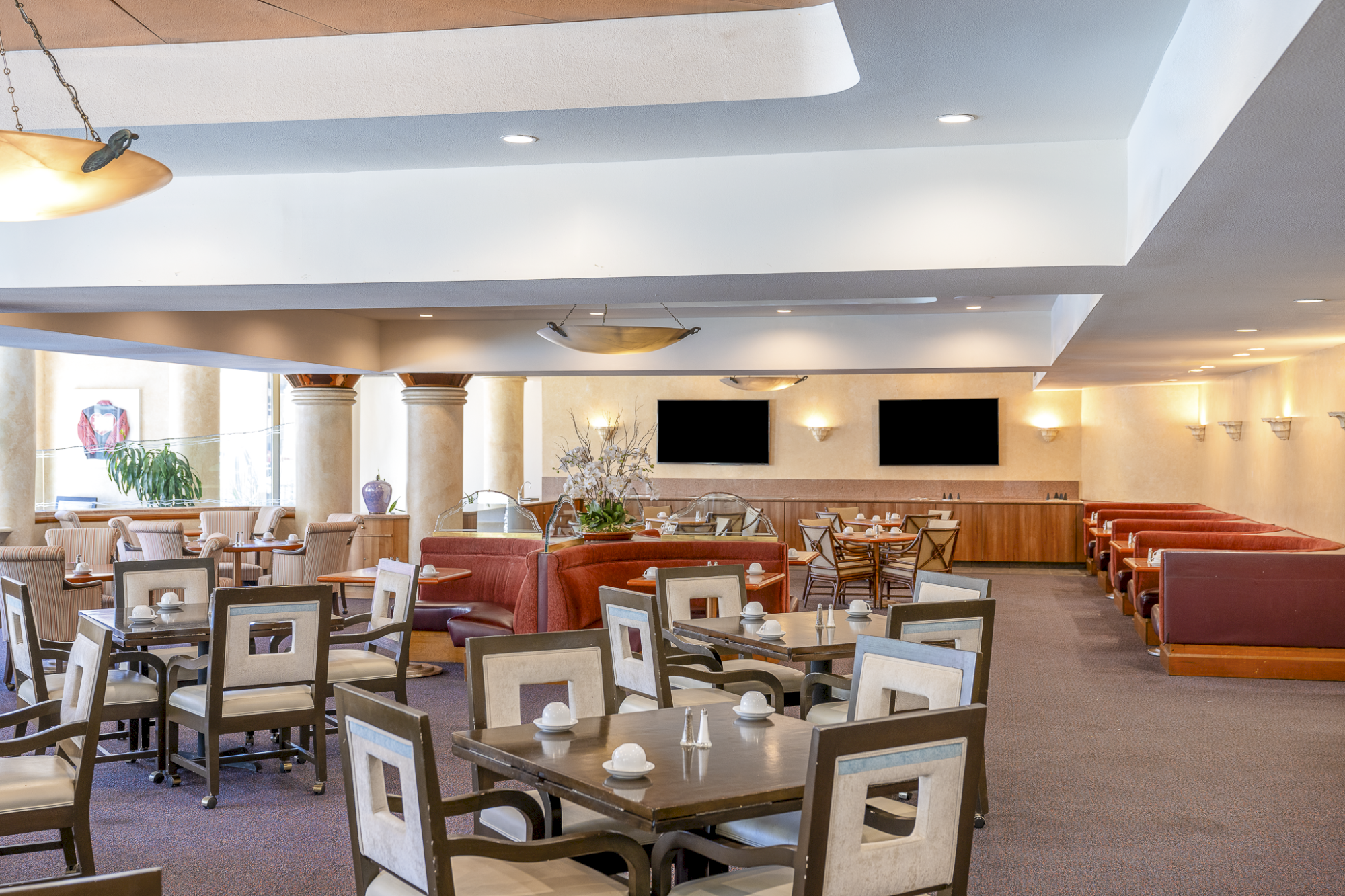 A spacious lounge or dining area with many tables, chairs, and seating, plus a bar and flat-screen TVs on the back wall, bathed in soft lighting.