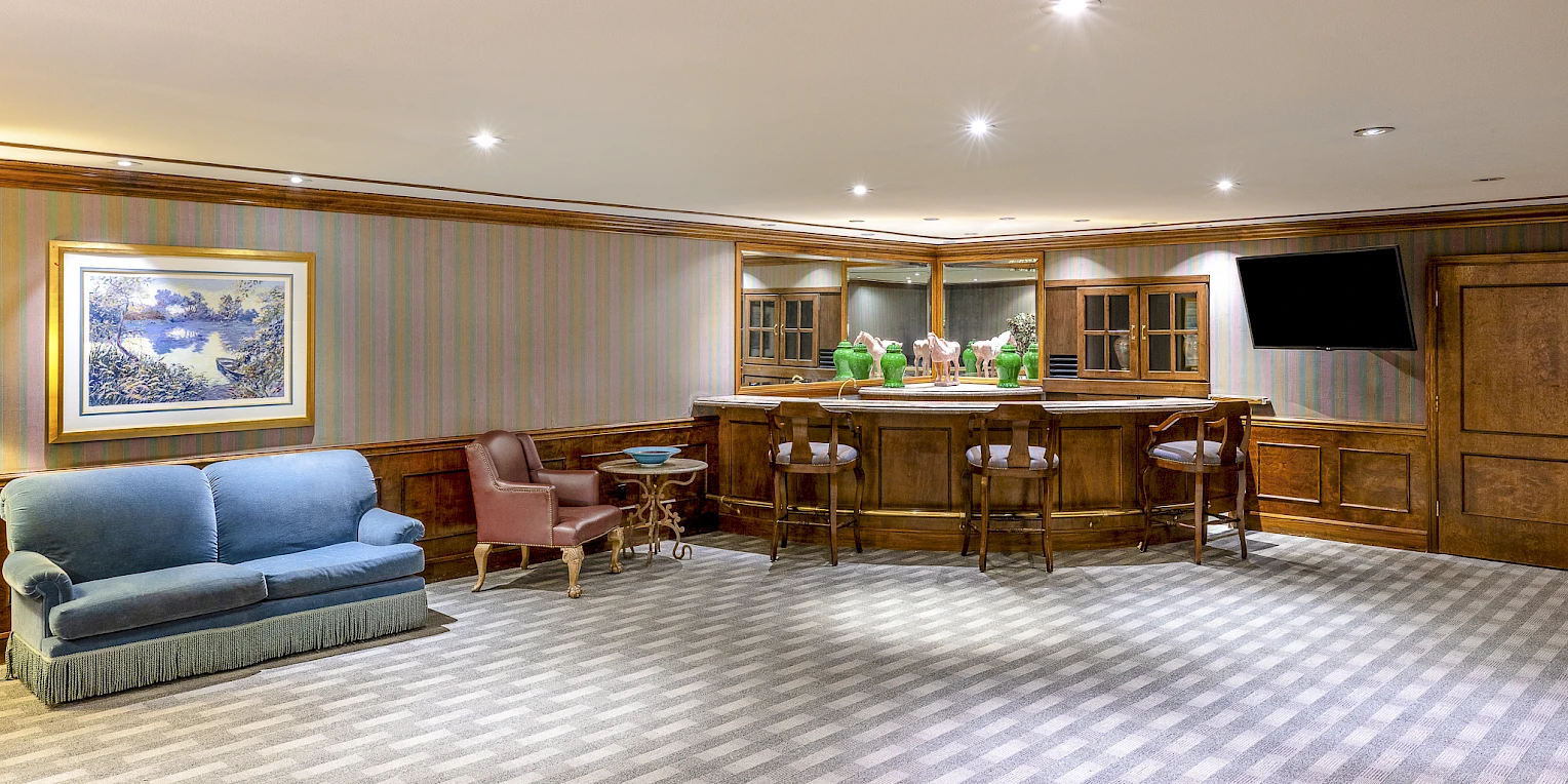 A cozy bar area with wooden paneling, a row of bar stools, a small seating area with a blue sofa and armchairs, framed art, and carpeted floor.