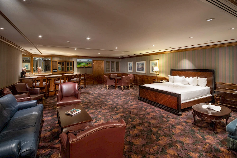 A spacious hotel suite with a plush bed, seating area, and wood accents; soft lighting, patterned carpet, and framed art along the walls.