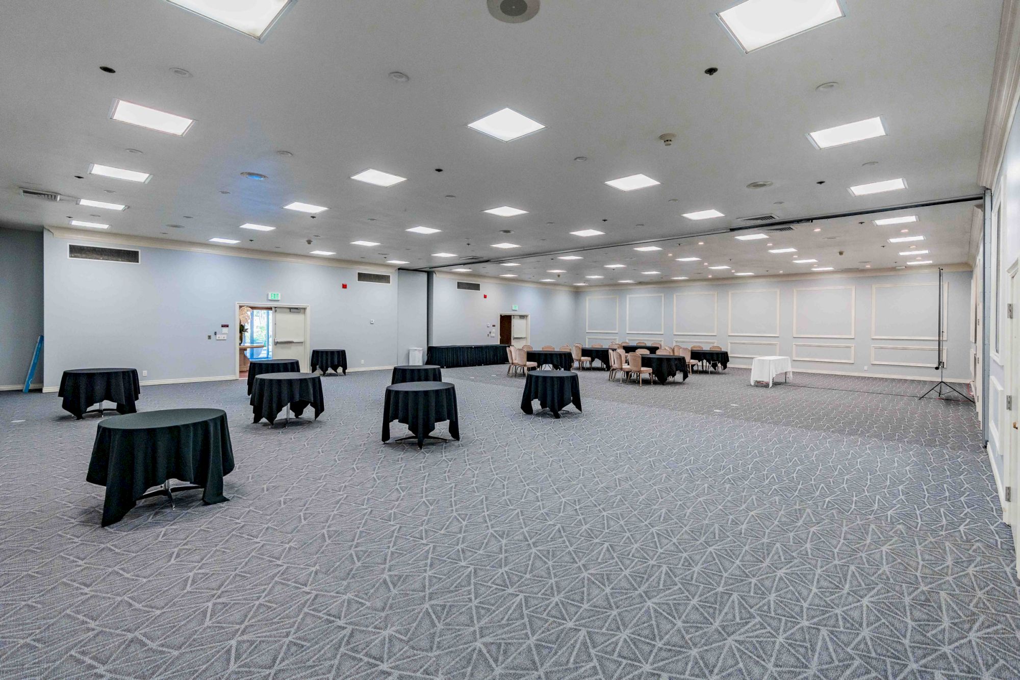 A large, empty event hall with high ceilings, blue-gray carpet, and several round tables covered in black cloth, spaced apart.