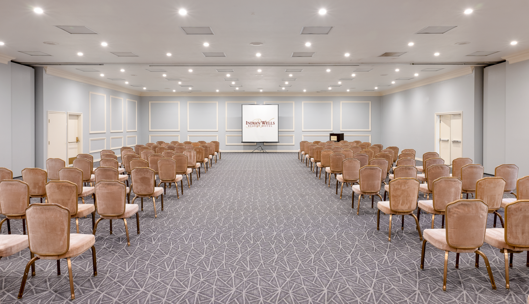 A large conference room with rows of brown chairs facing a stage and a projector screen at the front, soft blue walls, and bright ceiling lights.