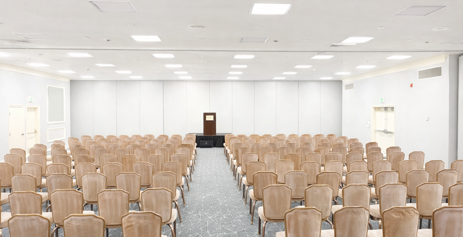 A large, empty conference room with many tan chairs arranged in rows facing a small stage podium at the front, bright overhead lighting.