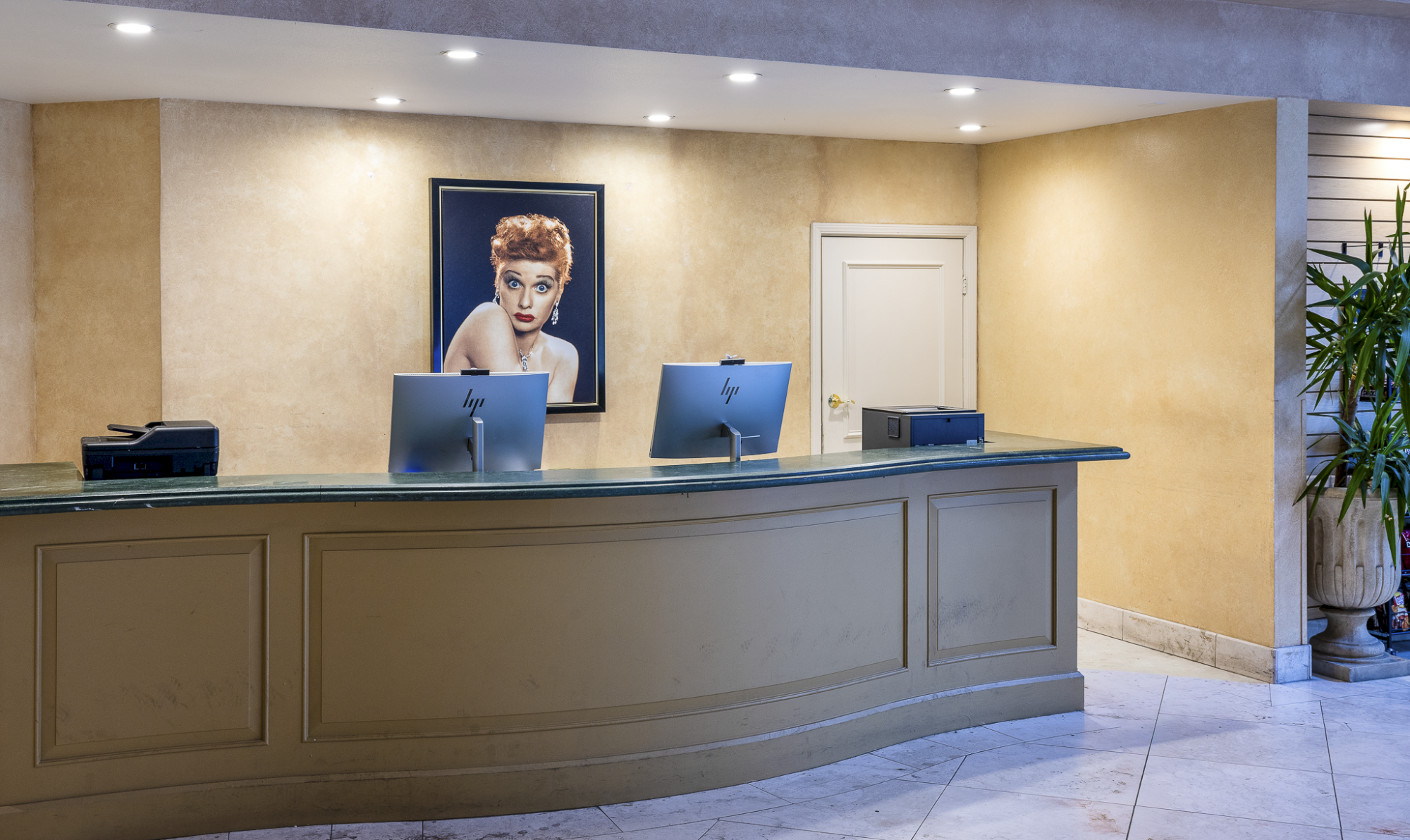 A hotel reception desk with two iMacs, a framed portrait on the wall, warm beige walls, a white door, and potted plant at right, quiet and tidy.