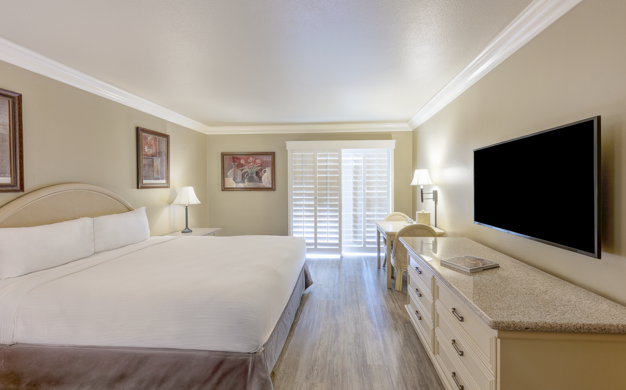 A hotel-style bedroom with a large bed, two nightstands, framed art, a dresser with a TV, and a desk by a window with shutters.
