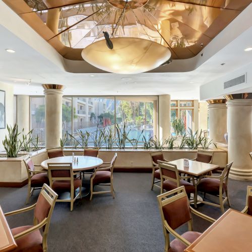 A bright, airy dining area with multiple tables and chairs, potted plants, large windows, and warm lighting. Top it at 140 characters, always ending the sentence.