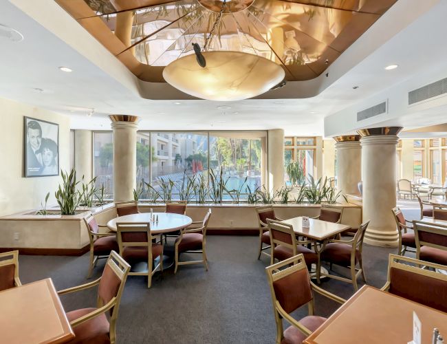 A bright, airy dining area with multiple tables and chairs, potted plants, large windows, and warm lighting. Top it at 140 characters, always ending the sentence.