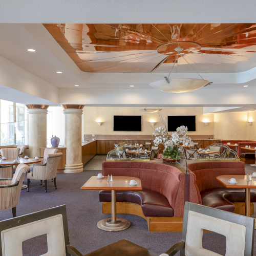 A spacious hotel lounge with cream chairs, red couches, small tables, plant decor, a bar area, and a glossy ceiling feature, bright and inviting.