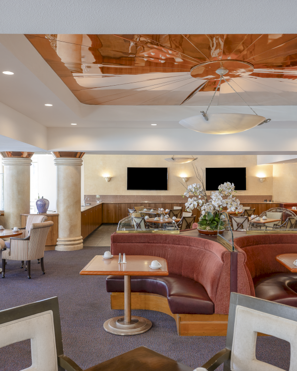 A spacious hotel lounge with cream chairs, red couches, small tables, plant decor, a bar area, and a glossy ceiling feature, bright and inviting.