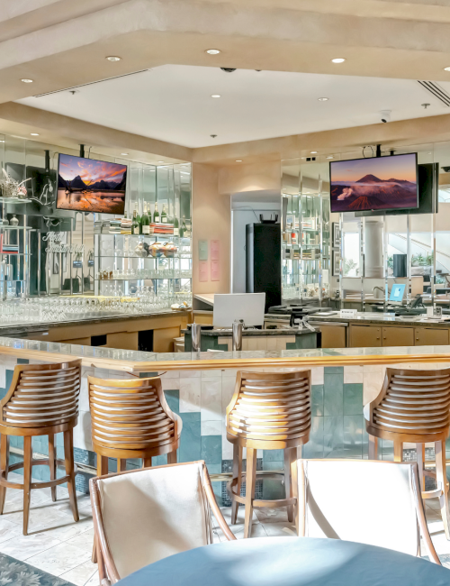 A modern cafe or bar with a curved counter, wooden bar stools, glass shelves, and multiple screens displaying scenic images, bright lighting, and a casual seating area.