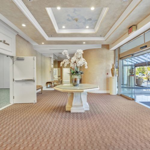A spacious hotel lobby with a central pedestal table, white flowers, warm lighting, and glass doors to the outside, cozy carpeted floor, and decorative ceiling.