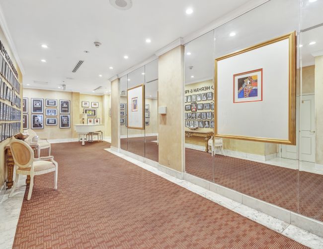 A neat gallery hallway with framed art on the walls, mirrors on the right, and a few chairs along the left; bright, tidy, and inviting. End.