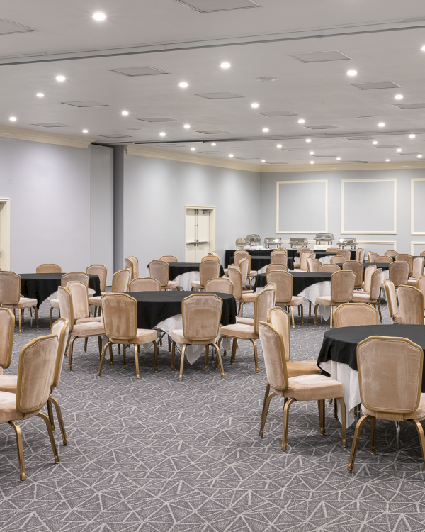 A large banquet hall with round tables draped in black cloths, beige chairs, and bright overhead lighting&mdash;ready for an event.