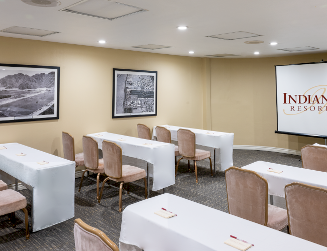 A conference room set up for a presentation, with rows of tables and chairs, a projector screen showing &ldquo;Indian Wells Resort Hotel,&rdquo; and framed photos on the wall.