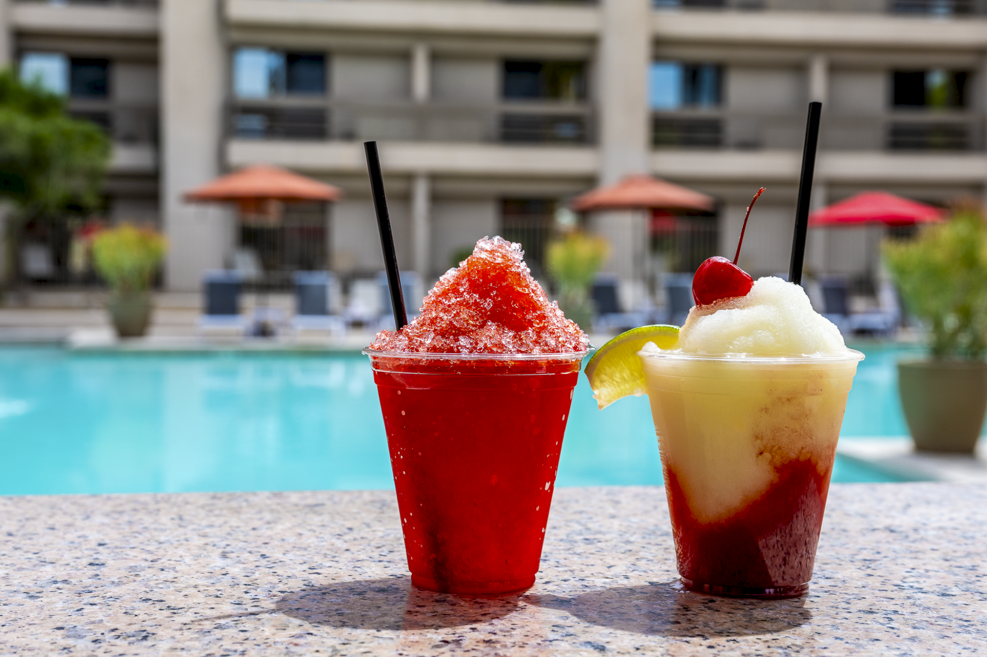 Two colorful frozen drinks on a poolside table with umbrellas in the background, one red slushy and one layered yellow-red with whipped cream.