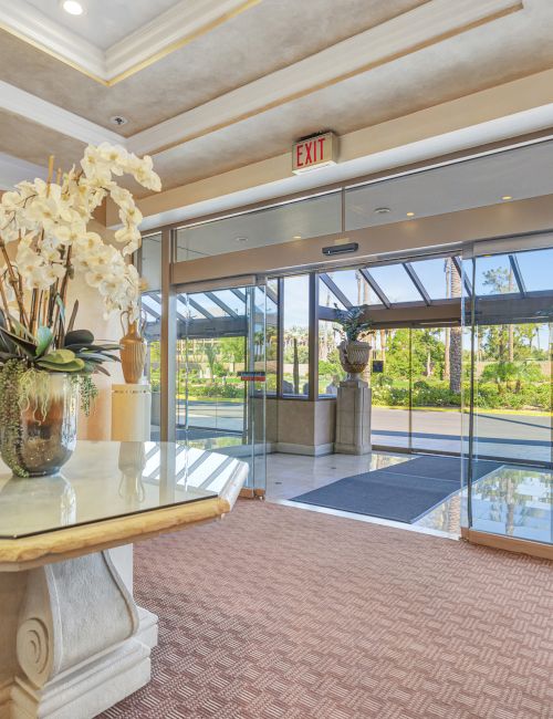 A hotel lobby with a marble pedestal table, decorative white flowers, large glass doors, and tasteful vases—sunlit and inviting.