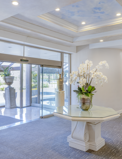 A modern hotel lobby with a glass entry, a central table holding a white orchid arrangement, soft grey carpet, recessed ceiling lights, and a bright, airy feel.