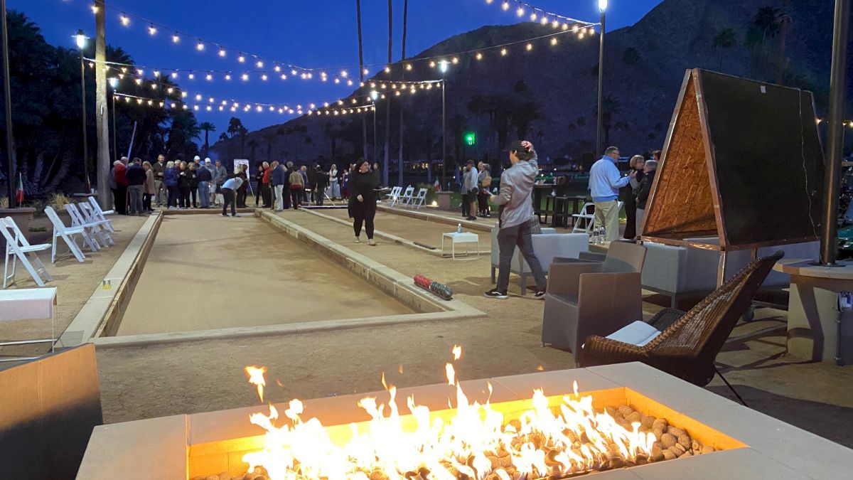 Beachside bocce court at dusk with string lights, fire pit, lounge chairs, and people gathering near the ramps.