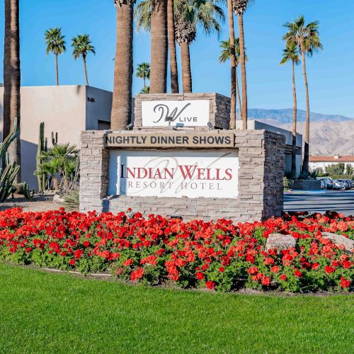 Sign reads “Indian Wells Resort Hotel” with a sign announcing nightly dinner shows; landscaped flower bed, palm trees, and mountains in the background.