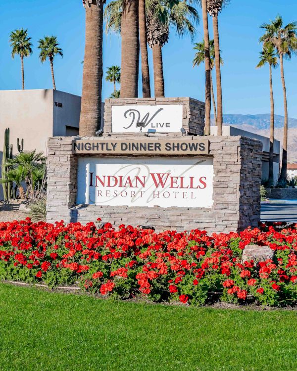 Sign reads “Indian Wells Resort Hotel” with a sign announcing nightly dinner shows; landscaped flower bed, palm trees, and mountains in the background.