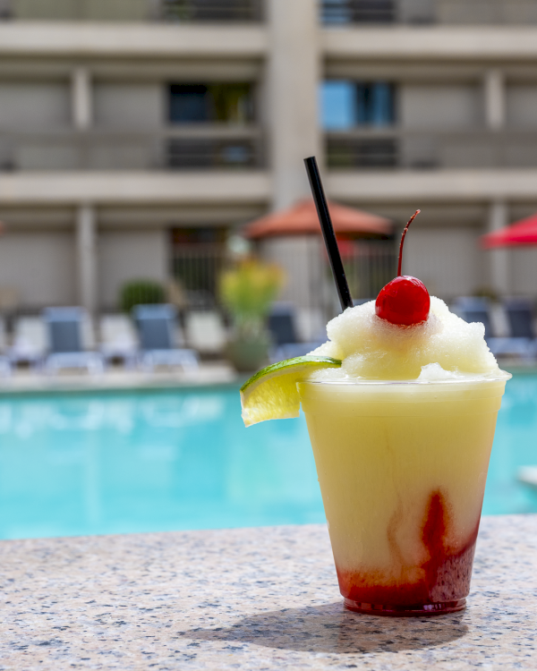 A tropical pineapple-strawberry smoothie with a cherry, lime wedge, and black straw on a poolside table by a sunny hotel pool.
