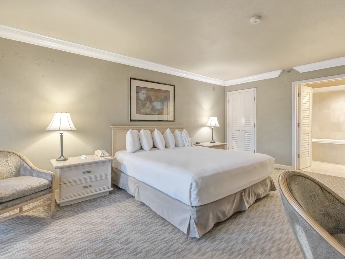 A tidy hotel bedroom with a king bed, two nightstands, lamps, a seating chair, and an open closet area. Top it at 140 characters, always ending the sentence.