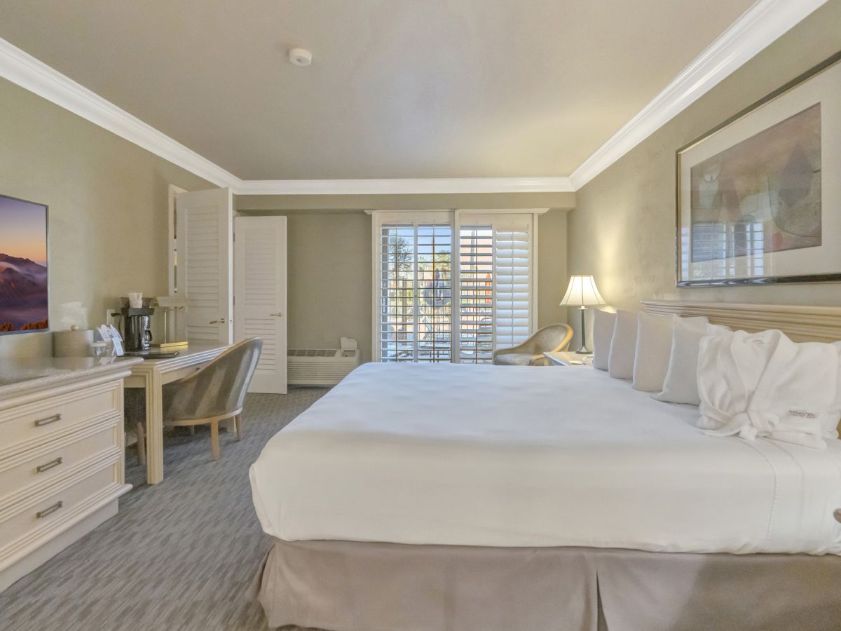 Hotel room with two queen beds, desk, chair, dresser, flat-screen TV, lamps, framed art, and large window with shutters. Ending with a period.