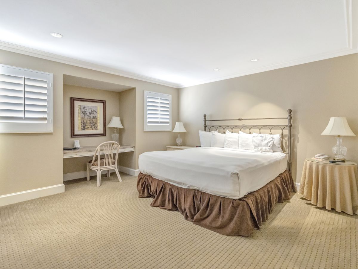 A bright hotel-style bedroom with a king bed, two nightstands, a desk and chair by the window, neutral walls, and beige carpet.
