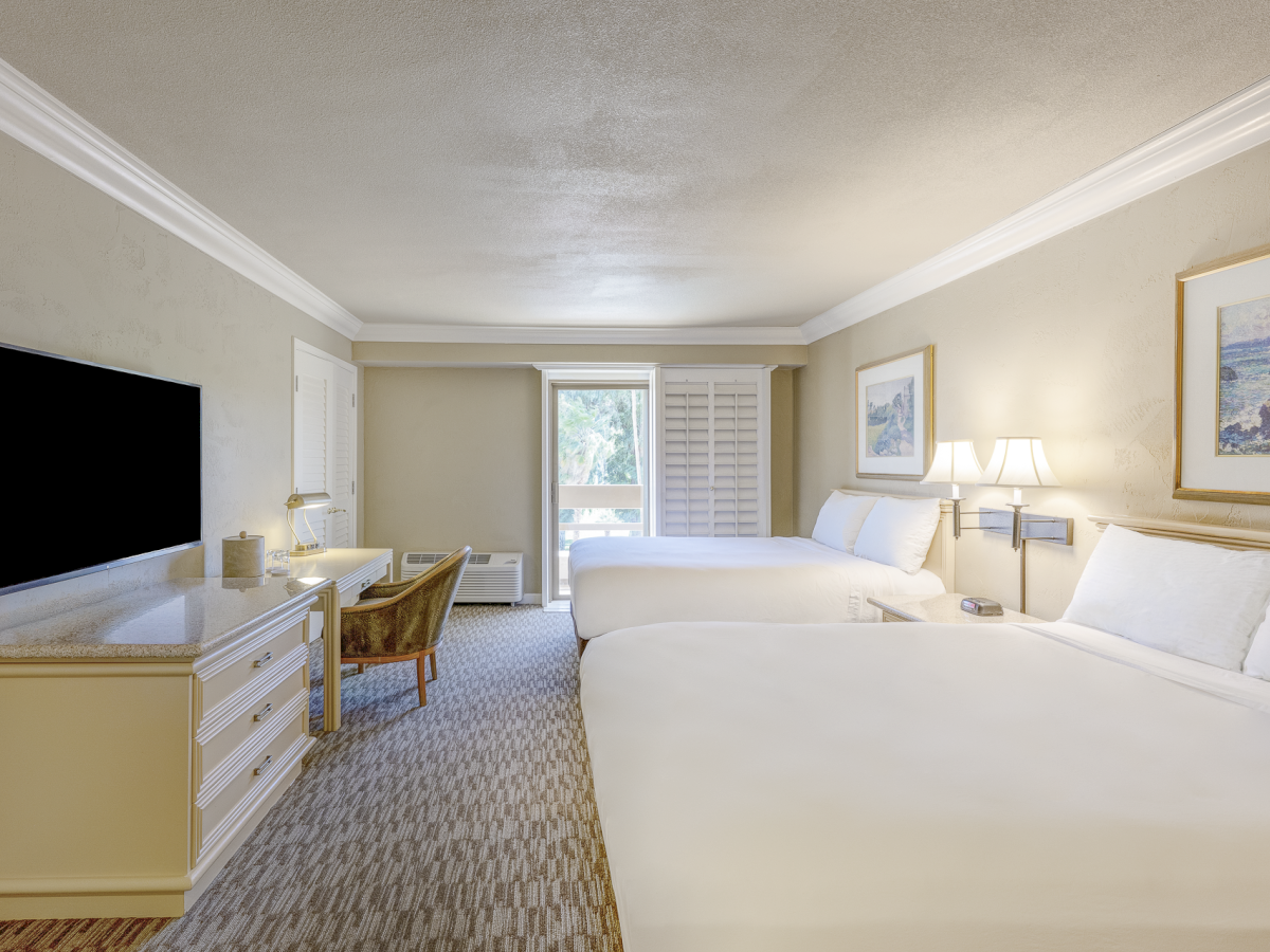 A hotel room with two large beds, a desk and chair, a TV on a dresser, a seating area, and a window with shutters, all in neutral tones.