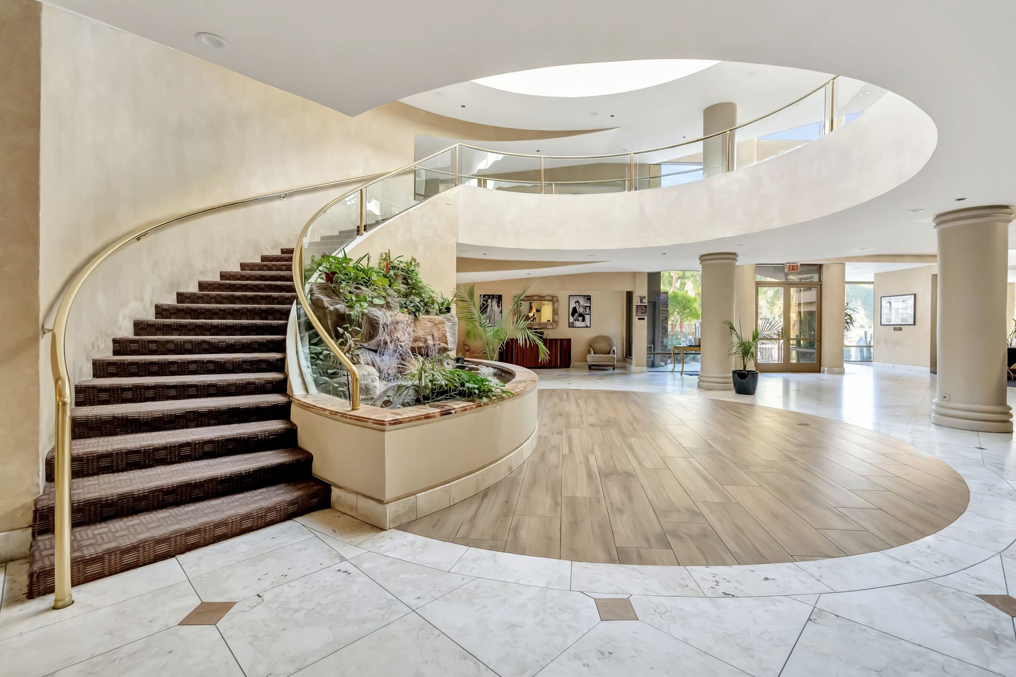 A grand hotel lobby with a curved staircase, circular balcony, modern decor, plants, and polished marble floors.