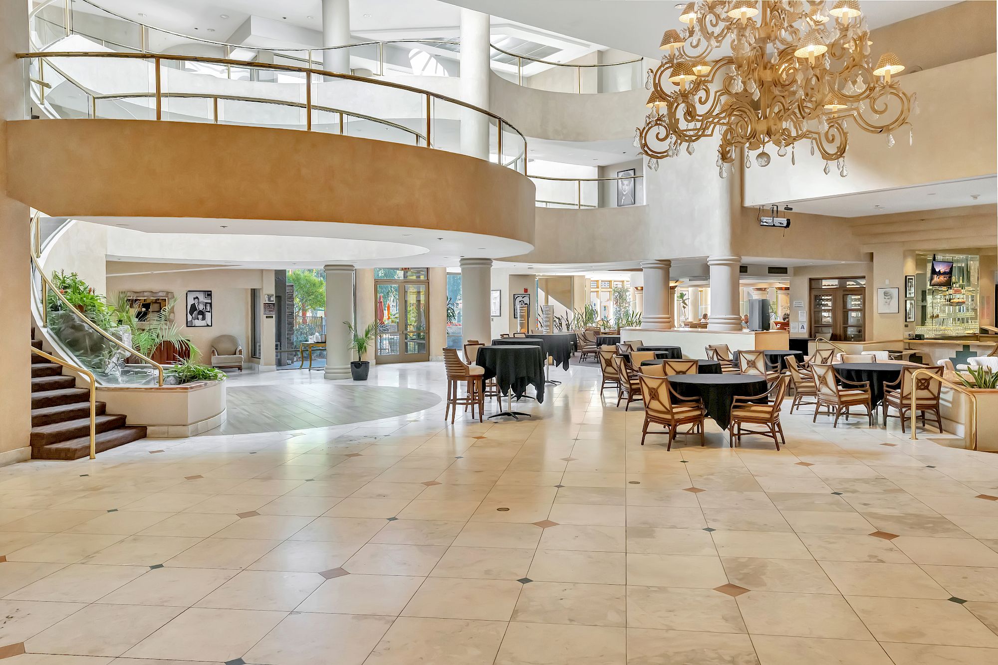 A spacious hotel lobby with a grand chandelier, curved staircase, marble floors, and several seating areas with tables and chairs. End with a period. 140 characters total.