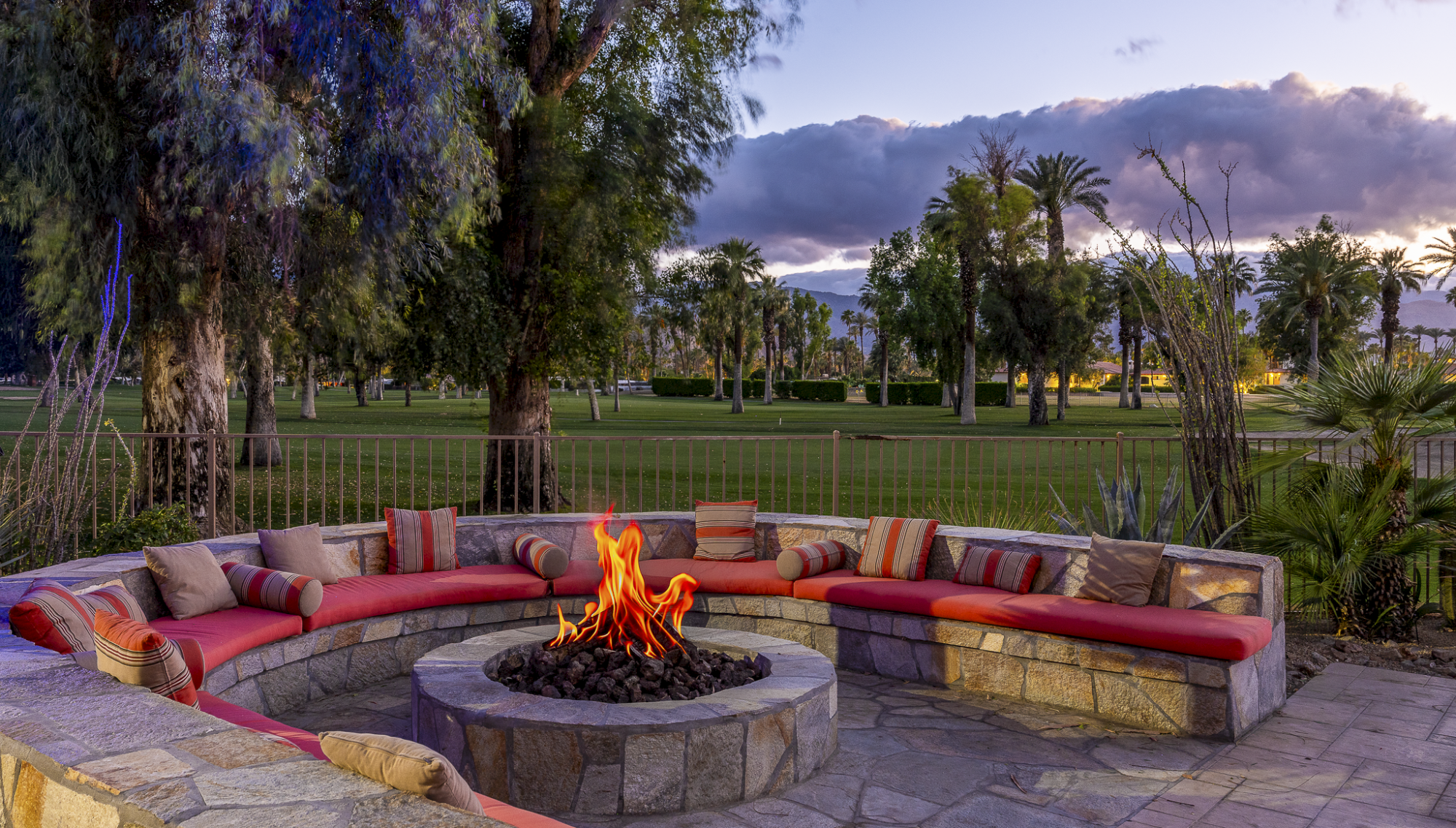 A circular stone seating area around a fire pit with a glowing flame, lush trees, palm trees, and a sunset sky in a park setting.