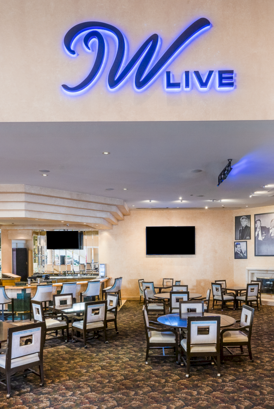 A hotel lounge or bar area with elegant seating, a blue &ldquo;WLIVE&rdquo; sign, warm lighting, and a TV screen on the wall, creating a sophisticated lounge vibe.