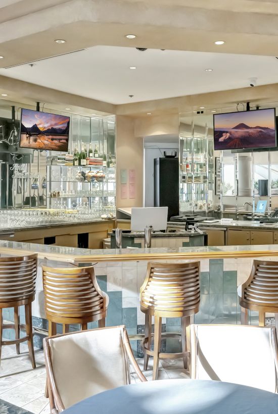 A bright hotel bar with a curved counter, wooden stools, wall-mounted TVs, framed photos, and a seating area in the foreground.