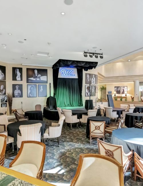 A spacious lounge or event room with vintage photos on the walls, a small stage with green curtains, and several round tables and chairs.