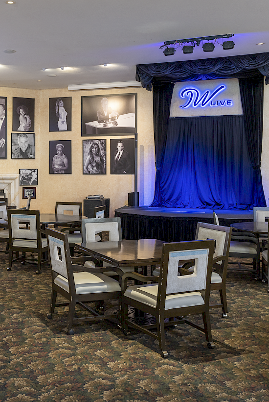A cozy lounge with a small stage, blue curtains, and framed photos on beige walls; chairs and tables arranged for intimate performances.
