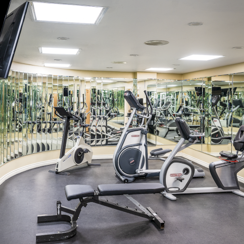 A brightly lit gym with treadmills and weight machines, mirrors on the walls, and people exercising in the background.