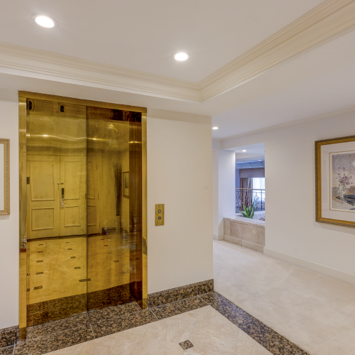 A warmly lit hallway with beige walls, framed art, and a wooden door at the end, finished with a light wooden floor and ceiling lights.