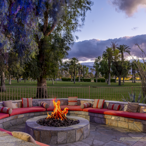 A sunny backyard with a circular fire pit, stylish seating, vibrant flowers, green lawn, and a scenic hillside view in the background.