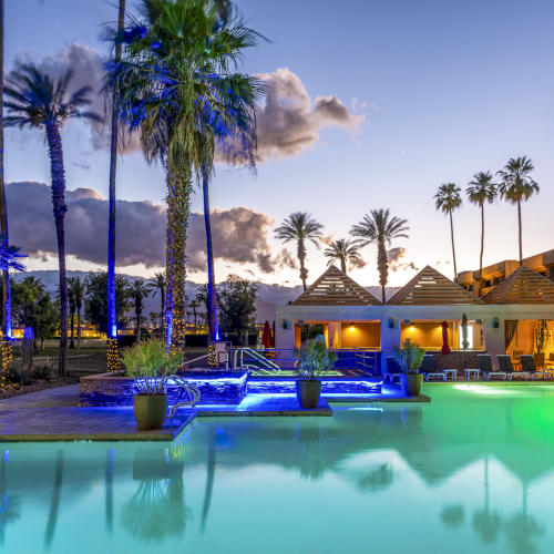 A luxurious resort pool at sunset with palm trees, lounge chairs, and modern architecture reflecting in the water.