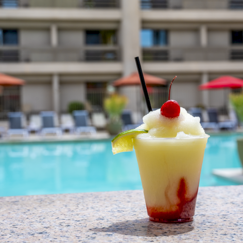 A tropical pool scene with a colorful drink in a glass, people lounging, and a hotel building in the background.