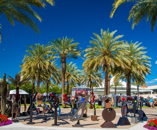 A sunny tropical street lined with palm trees, outdoor cafes, and people strolling along storefronts under a bright blue sky.