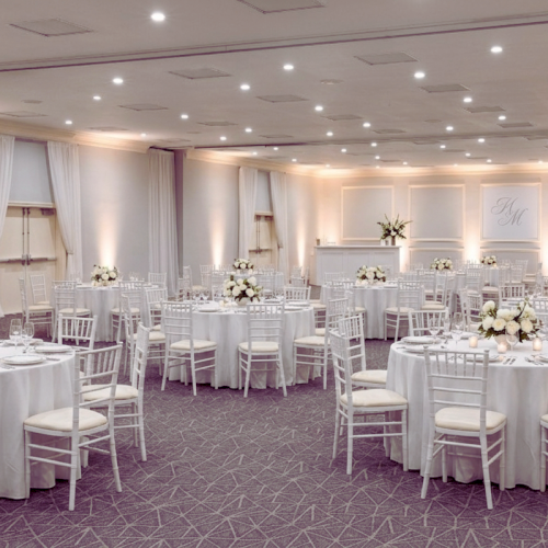 Elegant banquet hall with many round white-clothed tables, white chairs, soft lighting, and floral centerpieces&mdash;perfect for a formal event.