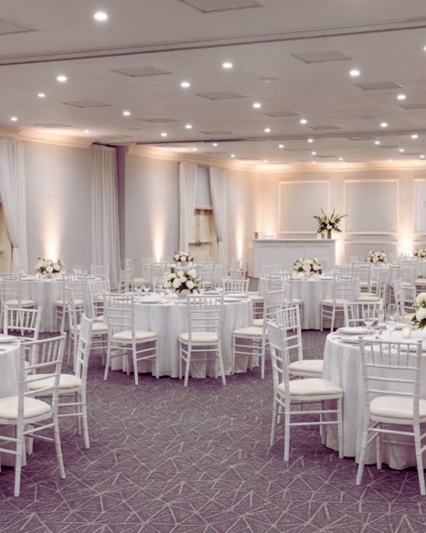 Elegant banquet hall with many round white-clothed tables, white chairs, soft lighting, and floral centerpieces&mdash;perfect for a formal event.