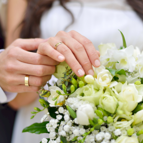A bride and groom hold a white bouquet; focus on the couple&rsquo;s hands with wedding rings, soft florals, and greenery in the bouquet.