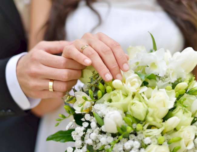 A bride and groom hold a white bouquet; focus on the couple&rsquo;s hands with wedding rings, soft florals, and greenery in the bouquet.