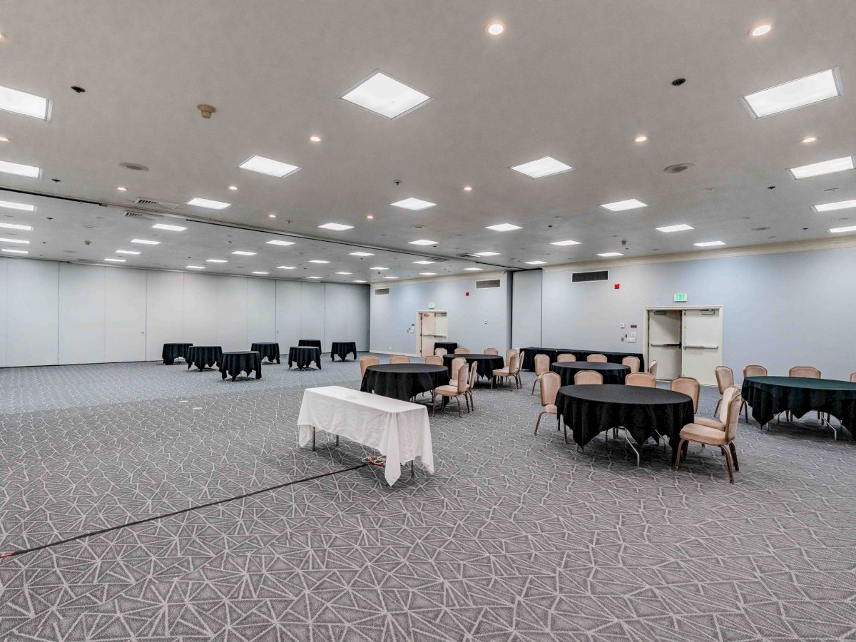 A large, empty conference room with multiple tables and chairs arranged in clusters, neutral walls, and bright overhead lighting.