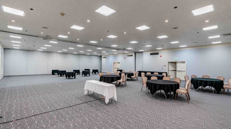 A large, empty conference room with multiple tables and chairs arranged in clusters, neutral walls, and bright overhead lighting.