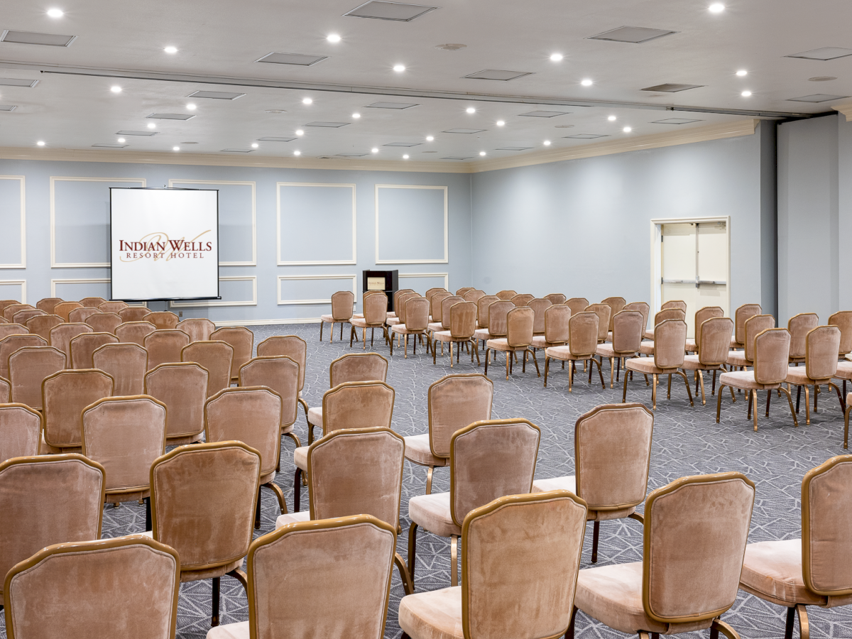 A large conference room set for a seminar, with many brown cushioned chairs facing a stage and screen at the front.