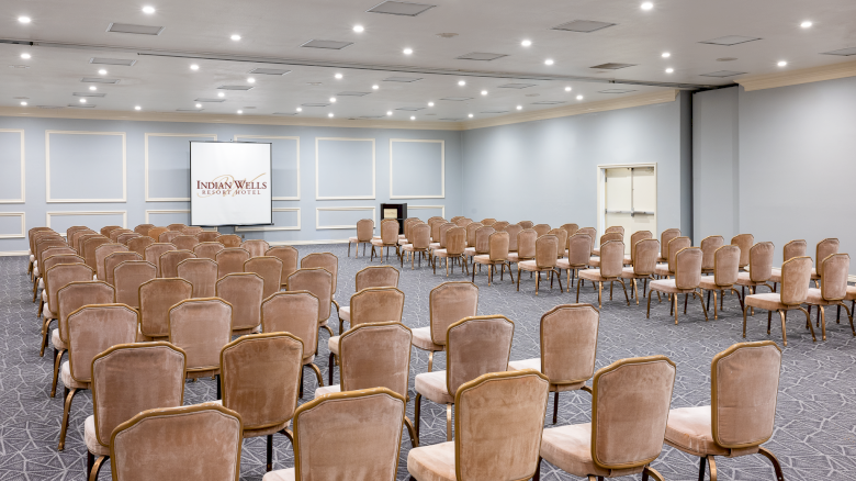A large conference room set for a seminar, with many brown cushioned chairs facing a stage and screen at the front.